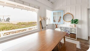 A room with a large window overlooking a beach, a wooden table, a blue cabinet, and a hanging mirror. - Carpinteria Beach Front Bungalow  (Carpinteria)