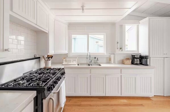 A kitchen with white cabinetry, a gas stove, a sink, and a window. - Carpinteria Beach Front Bungalow (Carpinteria)
