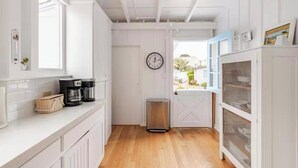 A narrow kitchen with white cabinetry, a coffee maker, a trash bin, and a clock on the wall. - Carpinteria Beach Front Bungalow  (Carpinteria)