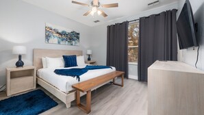 Ein modernes Schlafzimmer mit einem hölzernen Bett, Deckenventilator, einem großen Fenster mit Vorhängen, einem Nachttisch mit Lampe und einem an der Wand montierten Fernseher.