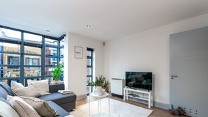 Un salon moderne avec un canapé gris, un téléviseur à écran plat posé sur un meuble blanc, une petite table blanche et une grande fenêtre donnant sur un immeuble.