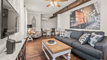 A living room with a grey sofa, wooden coffee table, and a ceiling fan. There is a dining area with a white table and chairs, and a large painting on the wall.