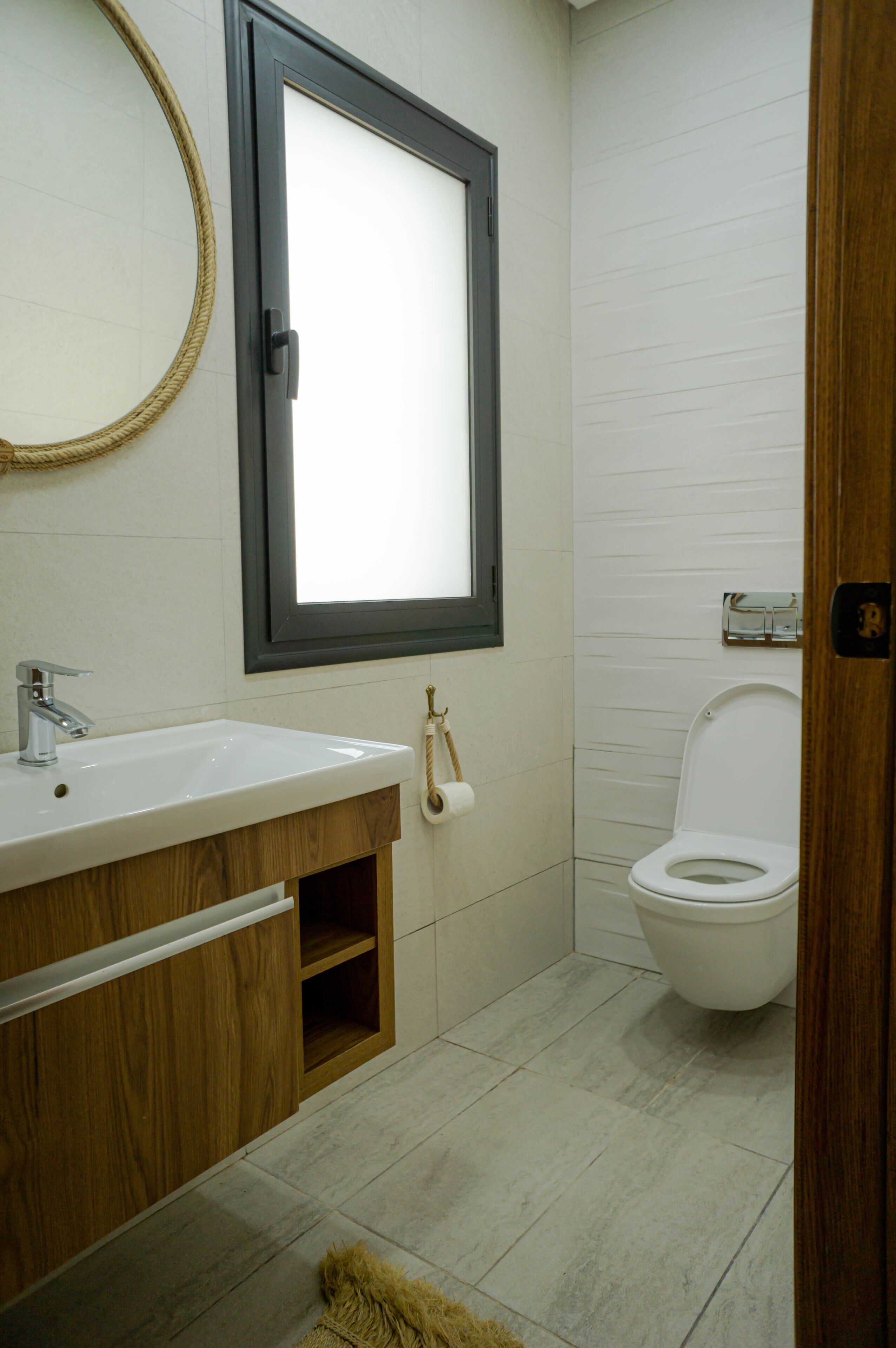 A bathroom with a wooden vanity, a round mirror, a toilet, and a small shelf.