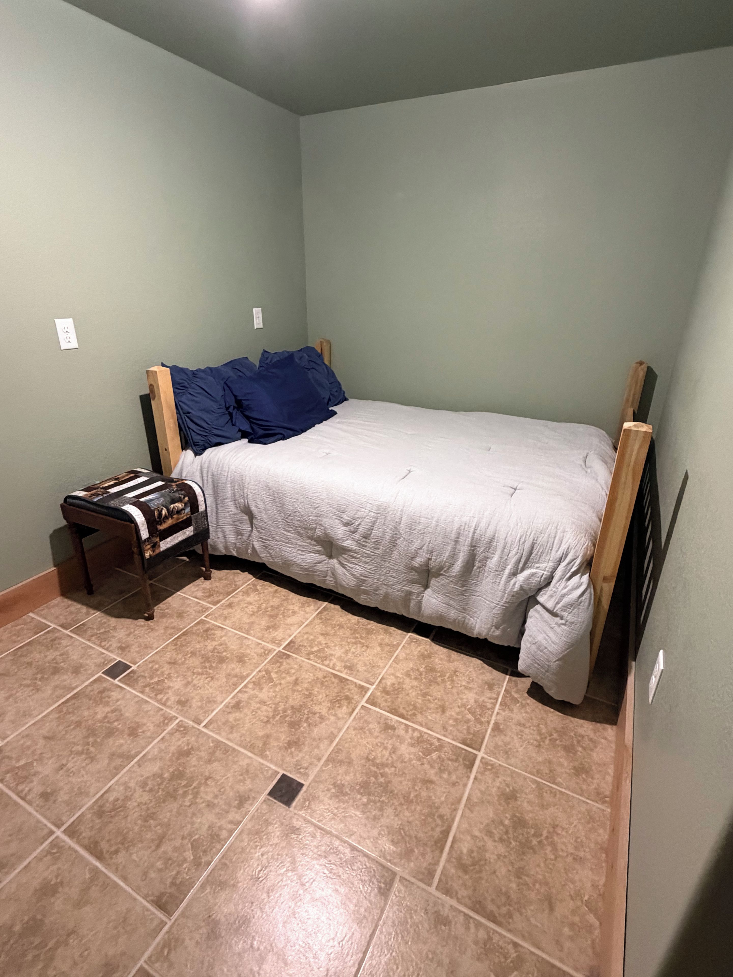 A small, single-bed room with a wooden headboard, a bedside table with a lamp, and a patterned bench.