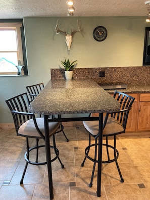 A kitchen with a granite countertop, a dining table with chairs, a potted plant, a wall-mounted clock, and a ceiling light fixture.