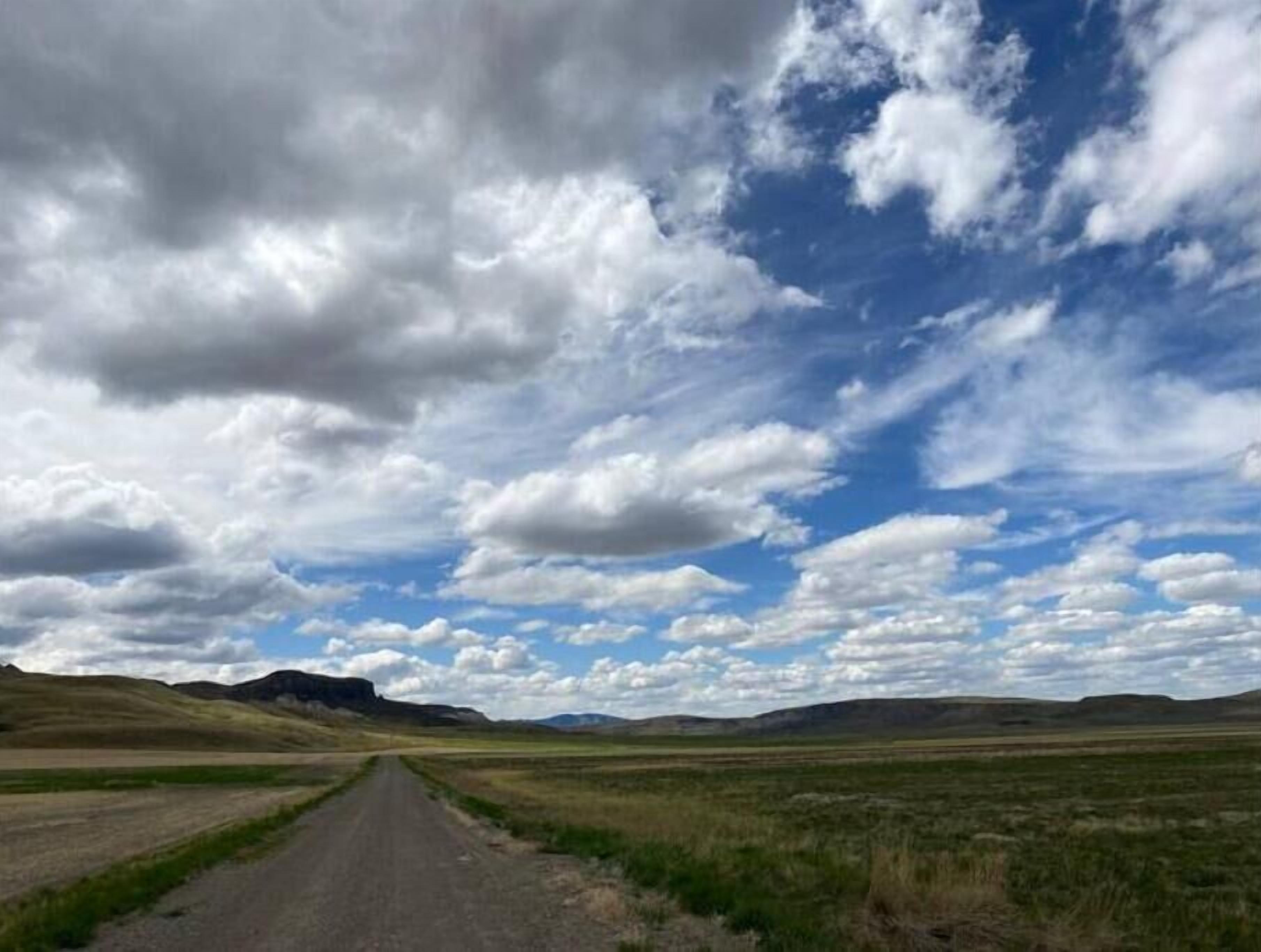 A rural road leading through a flat landscape with distant hills under a partly cloudy sky.