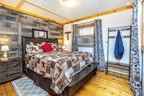 A bedroom with a wooden headboard, a quilted bedspread, a bedside table with a lamp, a bench, and a window with patterned curtains. - Cozy Ski Retreat on UTV/Snowmobile Trail (Merrillan)