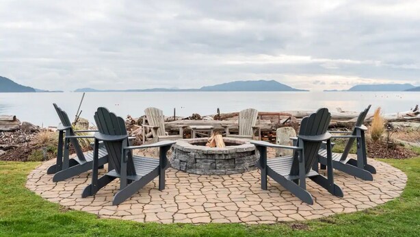 Un espacio de estar al aire libre con sillas Adirondack y una chimenea, con vistas a un lago y montañas al fondo.