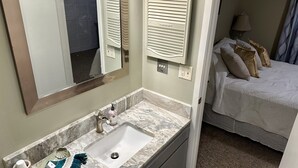 A bathroom with a marble sink, a mirror, and a toilet. There is a bed with pillows in the adjacent room. - 1st Floor 1BR/1BA Condo w/ Pool: Golf, Swim & Casino nearby! Sunshine Oaks (Diamondhead)