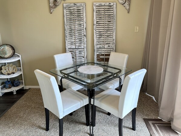 A dining area with a glass table surrounded by white chairs, a decorative plate on a wicker shelf, and two vintage-style shutters on the wall.