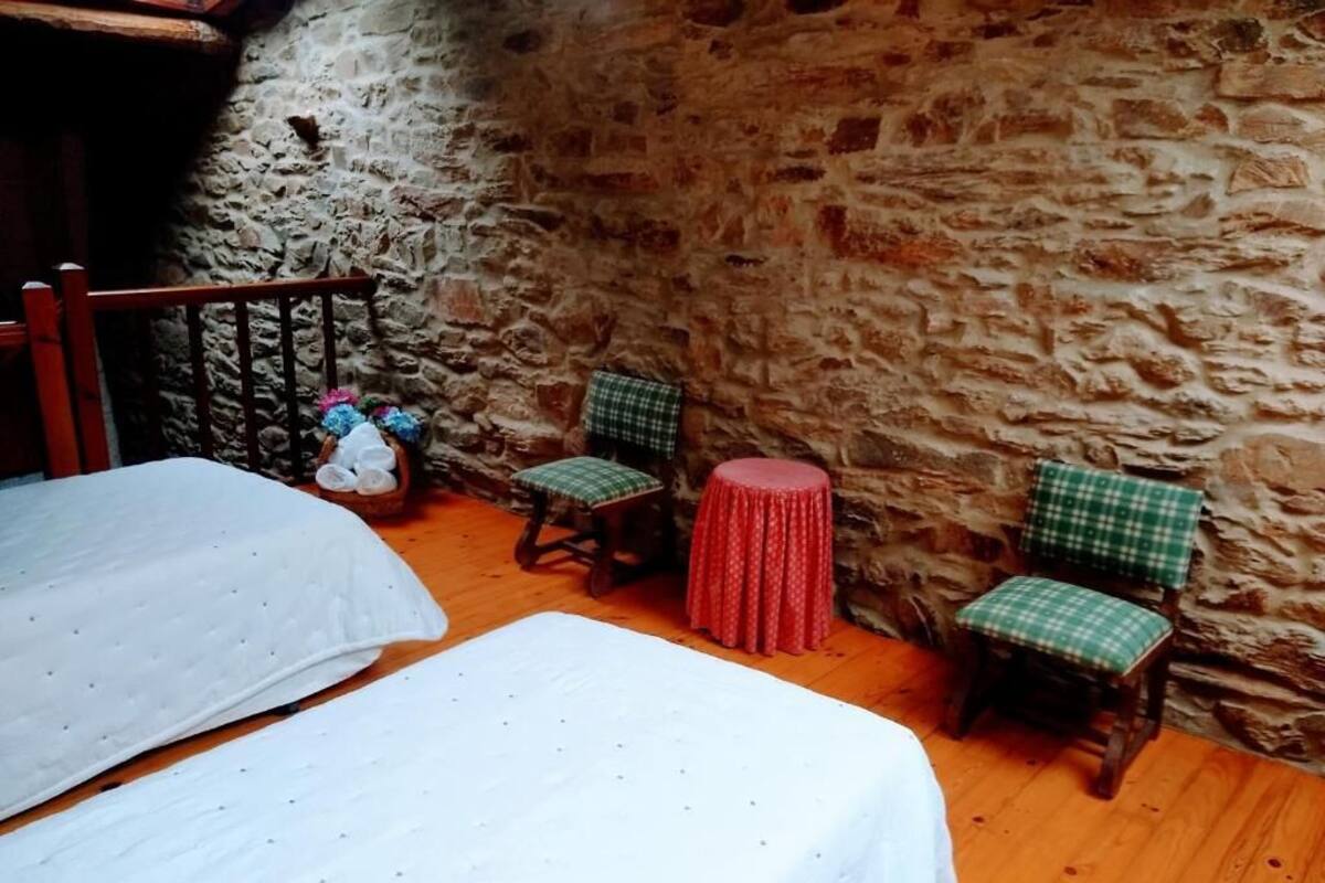 A rustic room with stone walls, wooden floors, and two beds with white linens. There are two green checkered chairs with red cushions and a small round table with a red tablecloth.