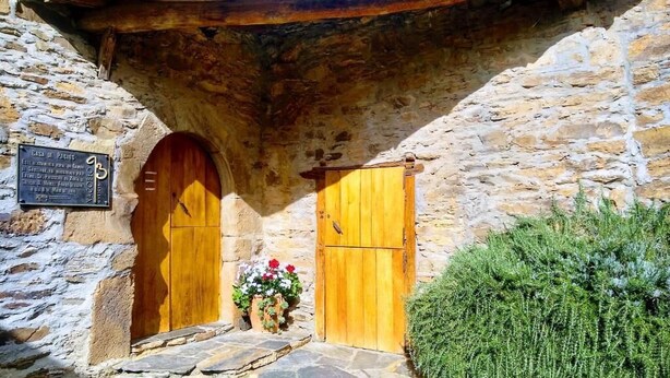 A stone building with two wooden doors, a small garden with flowers, and a sign on the wall. - Casa Pacios By M De Santiago (Triacastela)