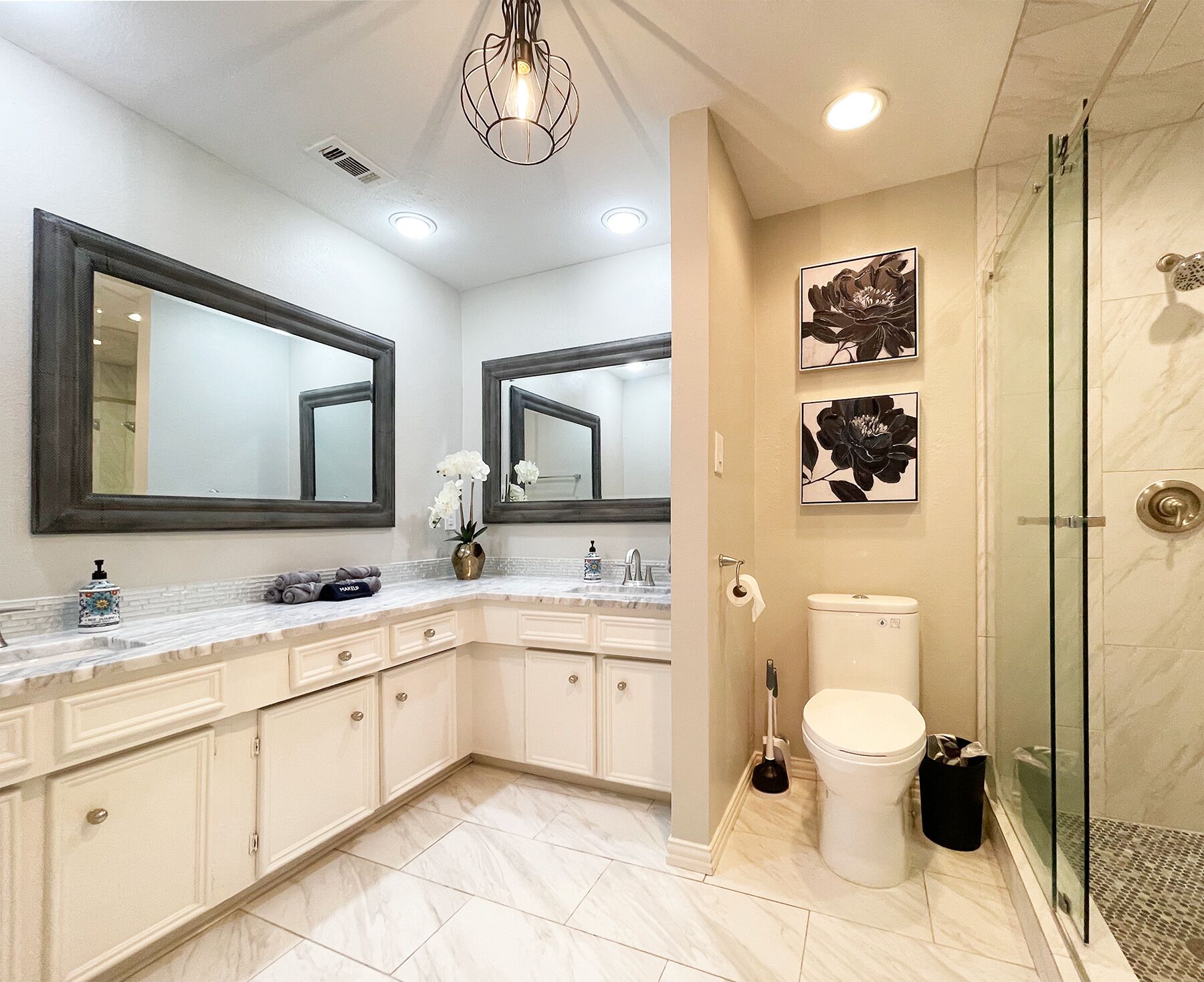 Een moderne badkamer met een marmeren wastafelmeubel, twee grote spiegels, een toilet, een douche met glazen deuren en kunst aan de muur.