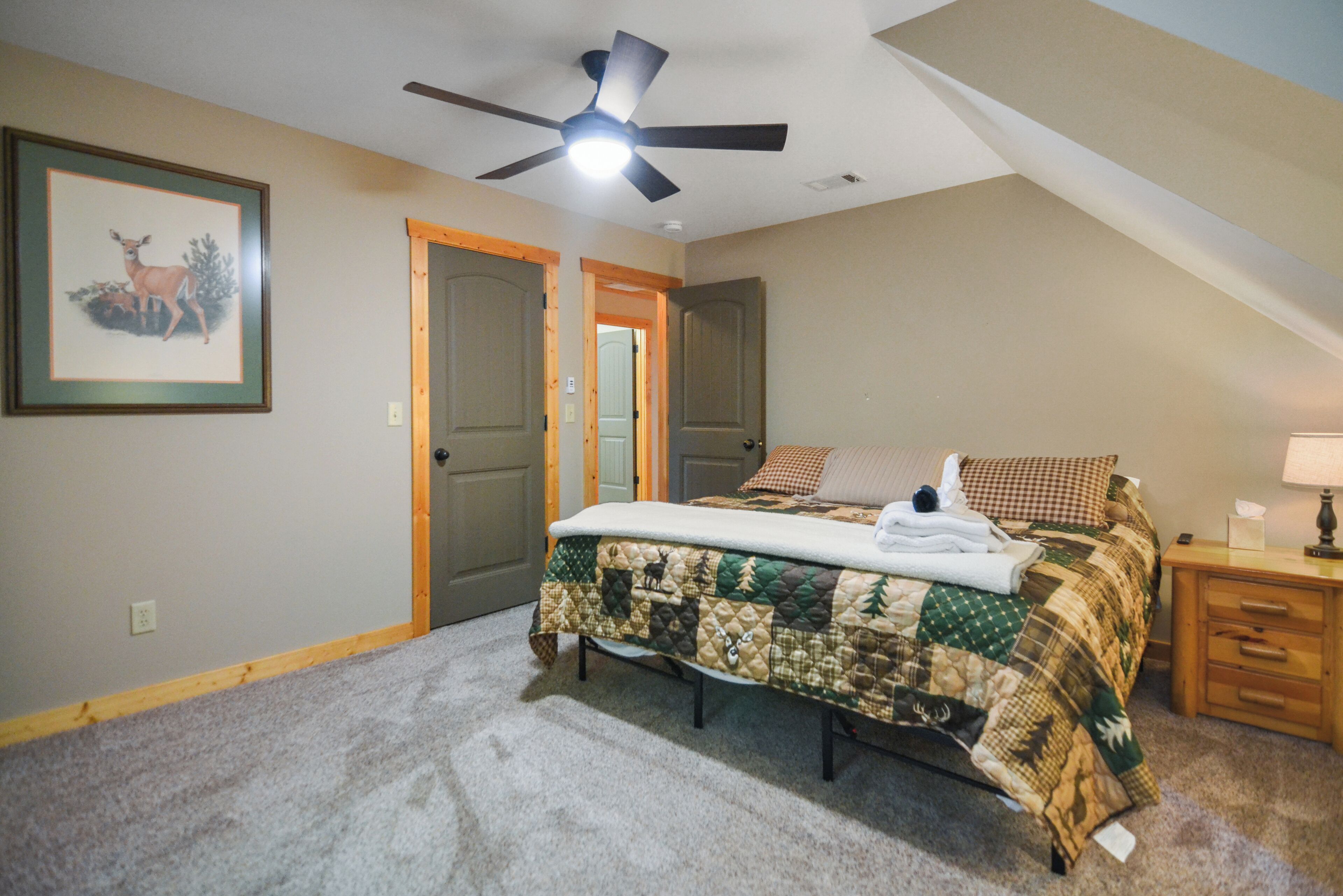 Une chambre à coucher avec un lit, un ventilateur de plafond, un tableau représentant un cerf encadré et une porte.