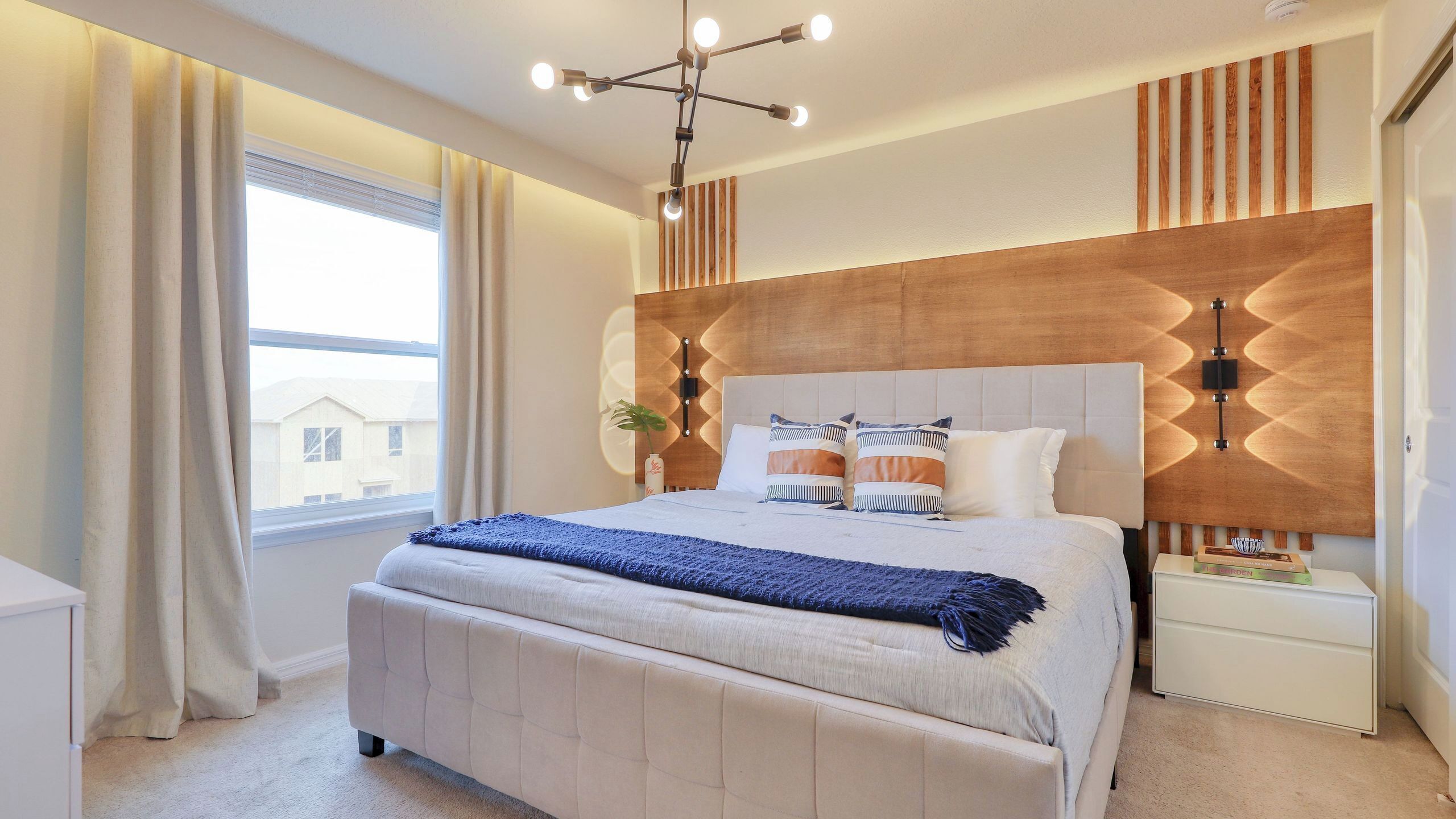 A bedroom with a large bed, bedside tables, a modern chandelier, and a view of buildings outside.