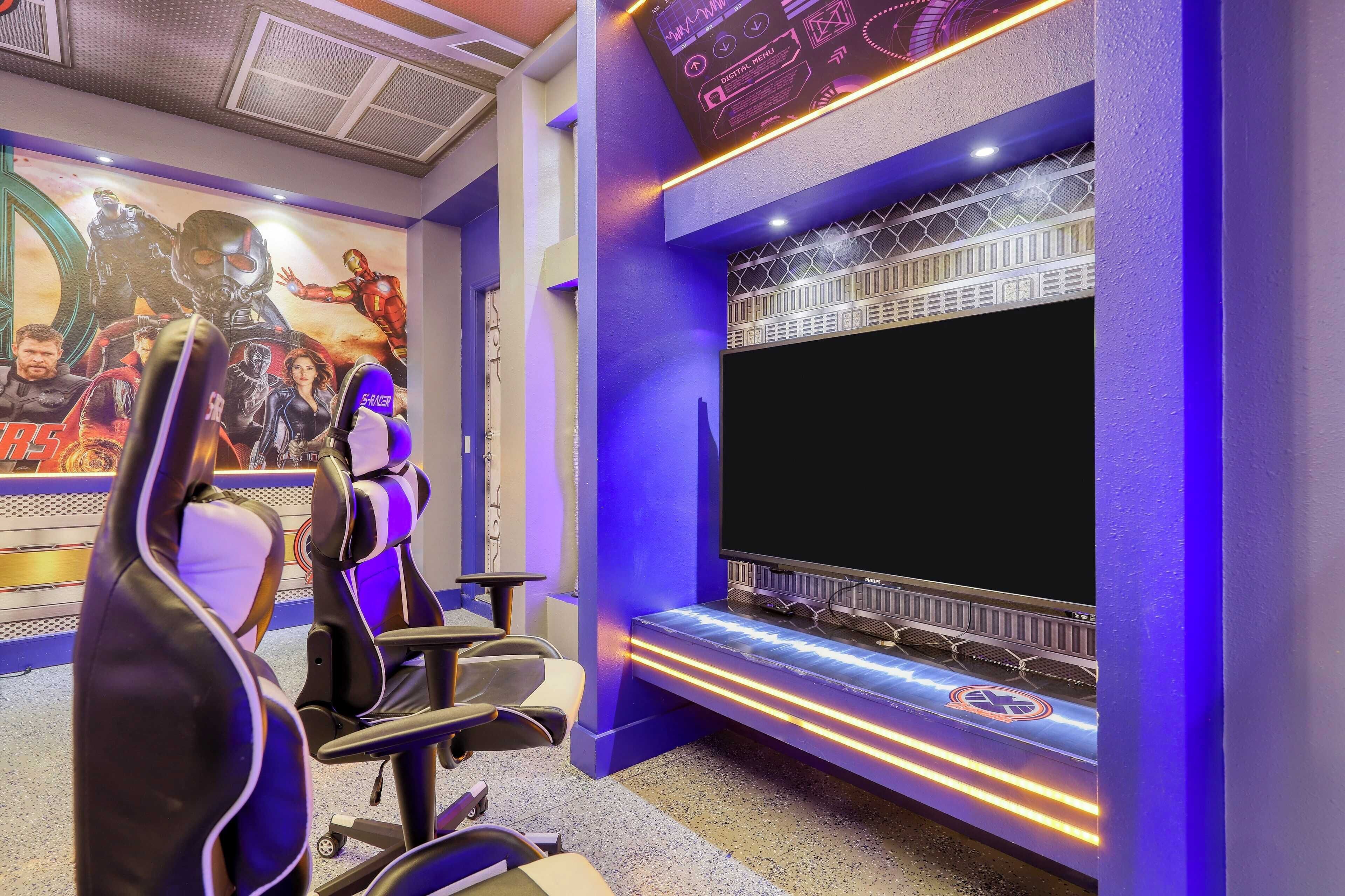 Una sala de juegos con una pantalla grande, sillas gamer y un mural de superhéroes en la pared.
