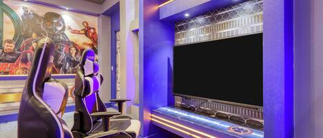 Una sala de juegos con una pantalla grande, sillas gamer y un mural de superhéroes en la pared.