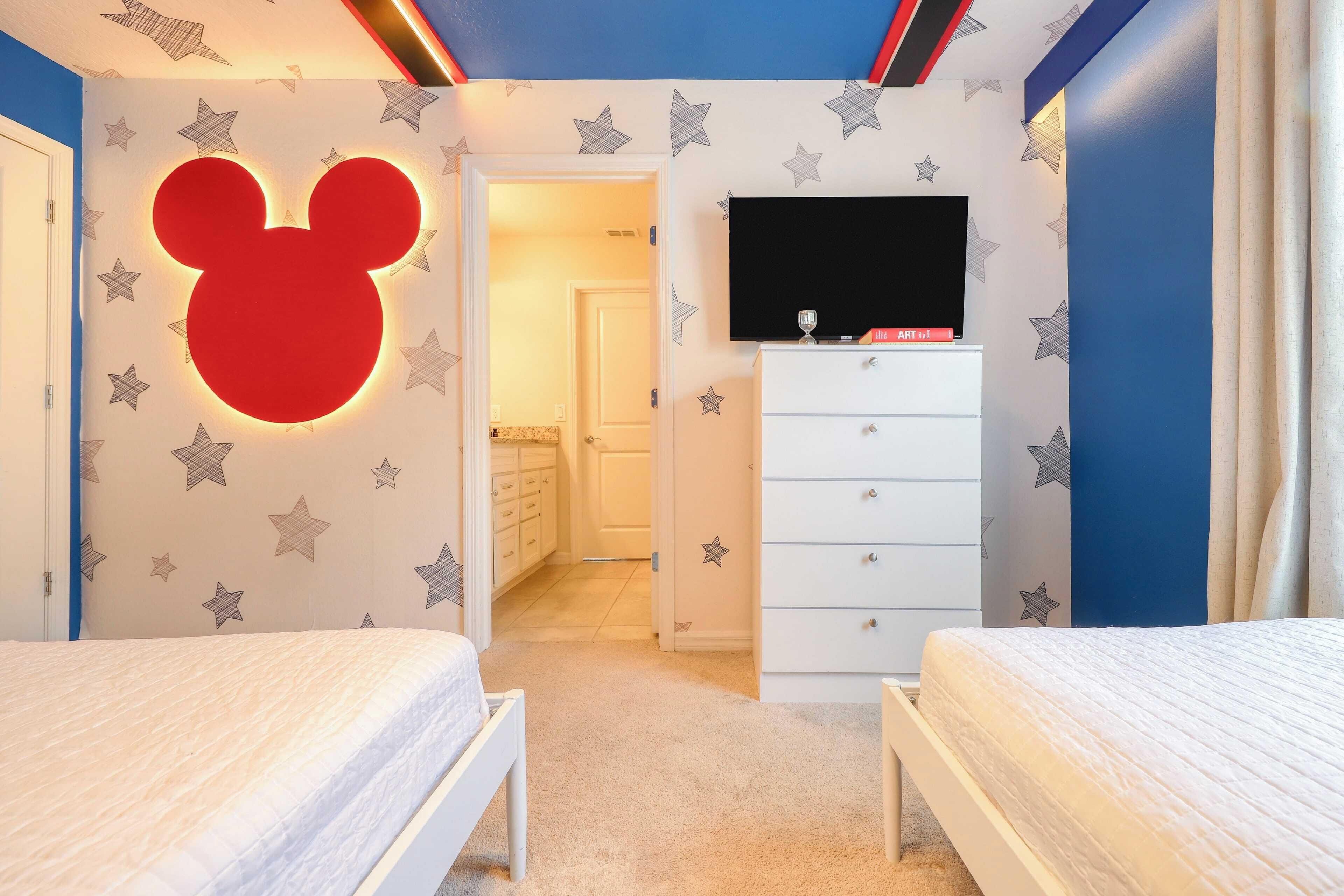A child's bedroom with a large Mickey Mouse decal on the wall, a TV, a dresser, and two beds.