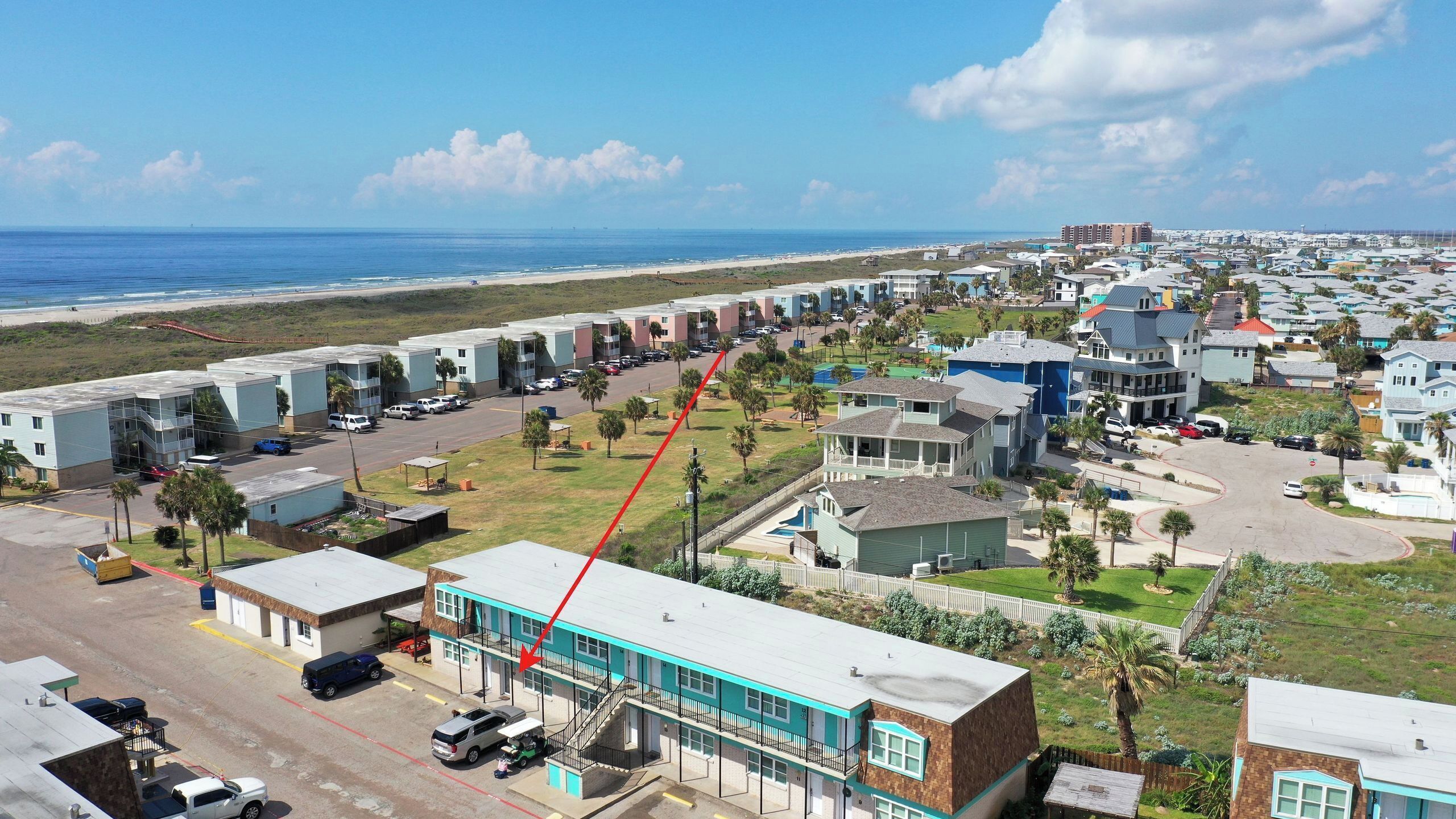 Een woongebied aan de kust met een rij appartementen aan het strand, een parkeerplaats en een klein gebouw met een blauw dak.