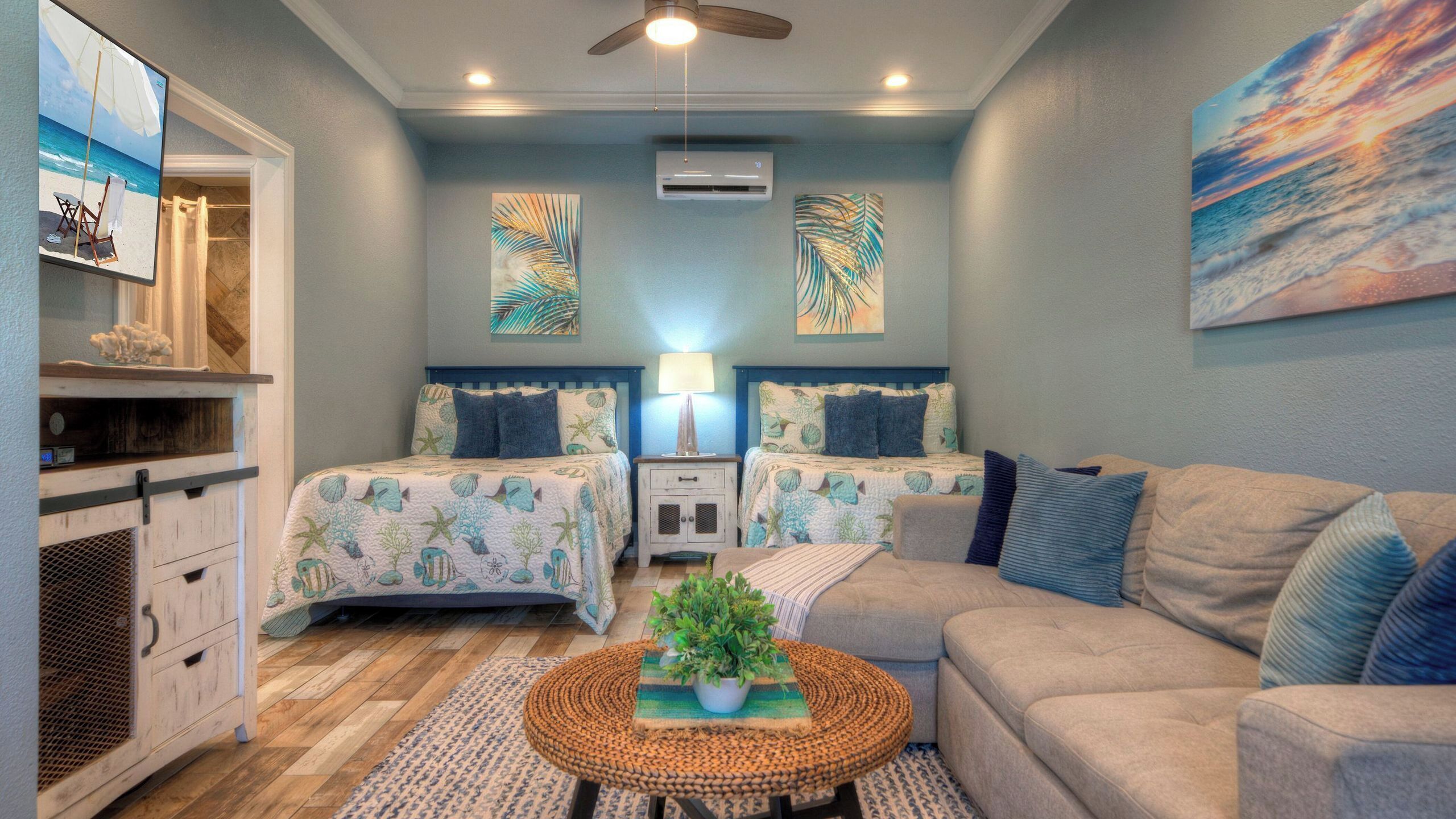 A room with two beds, a sofa, a ceiling fan, an air conditioning unit, and a beach-themed artwork.