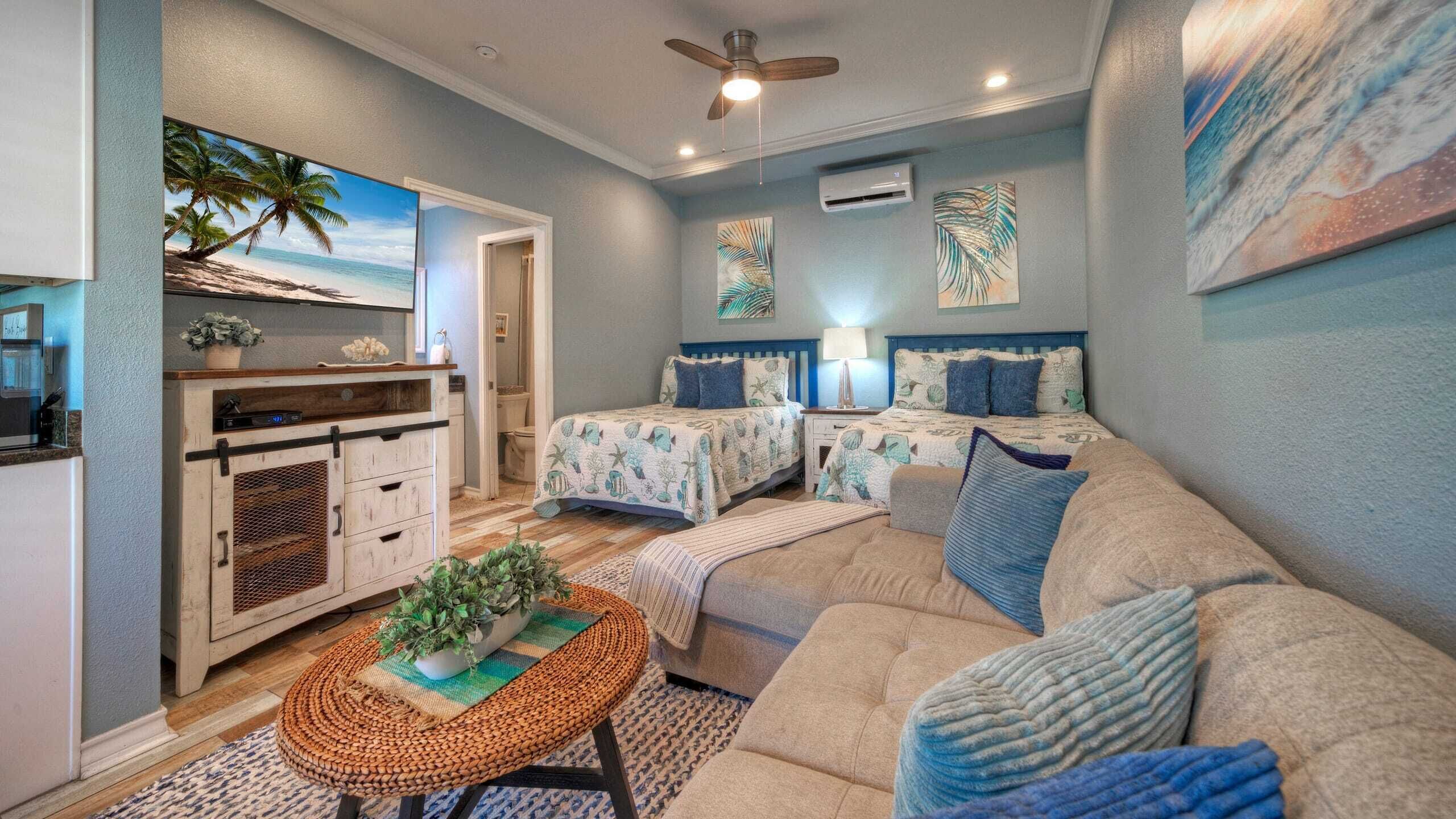 A room with a kitchenette, two beds with matching headboards, a ceiling fan, and artwork depicting tropical scenes.