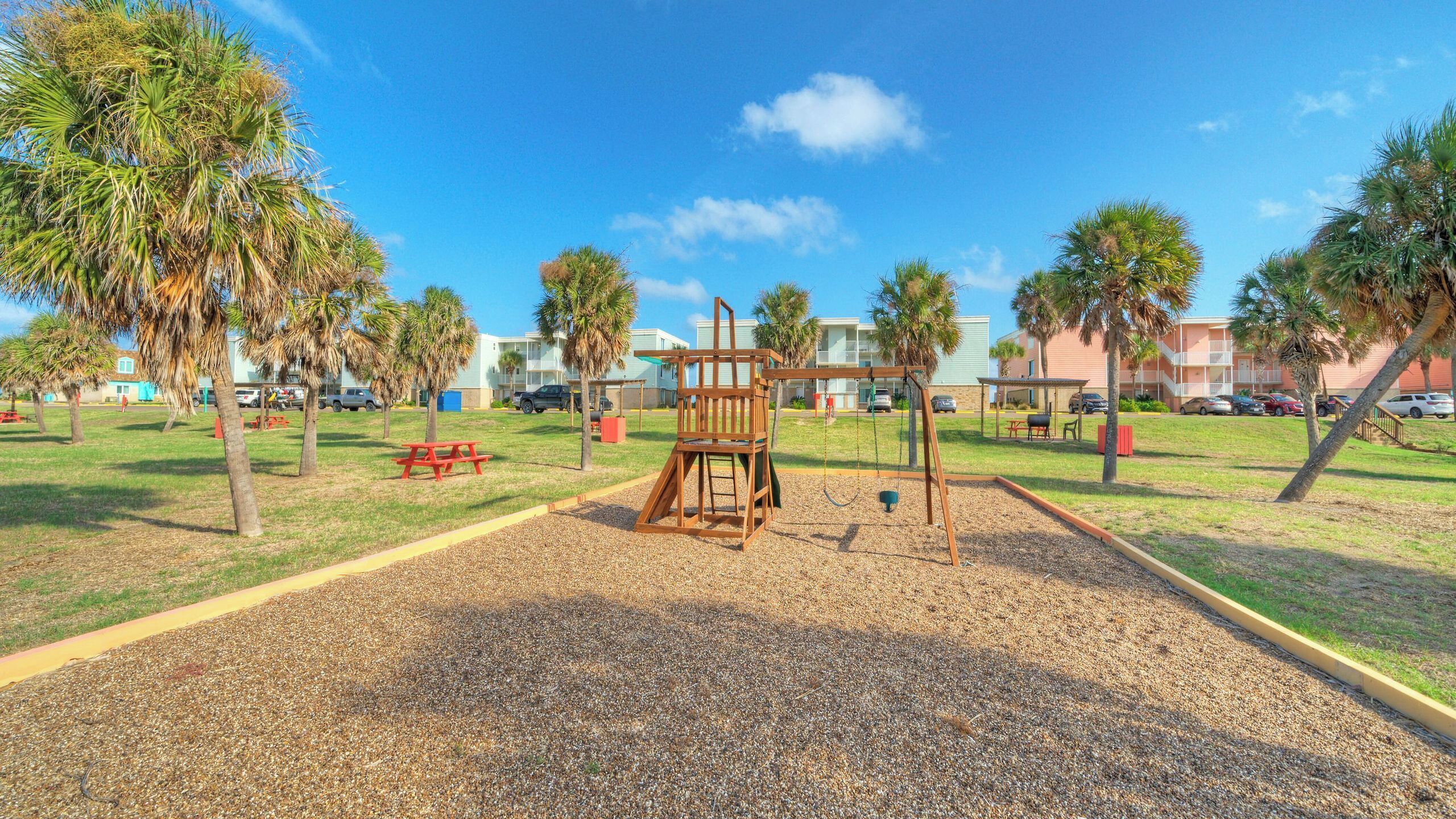 A playground with a wooden swing set, a red picnic table, and palm trees.