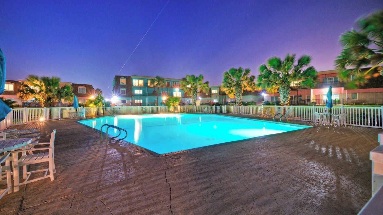 A swimming pool with a wooden deck, palm trees, and a building in the background.