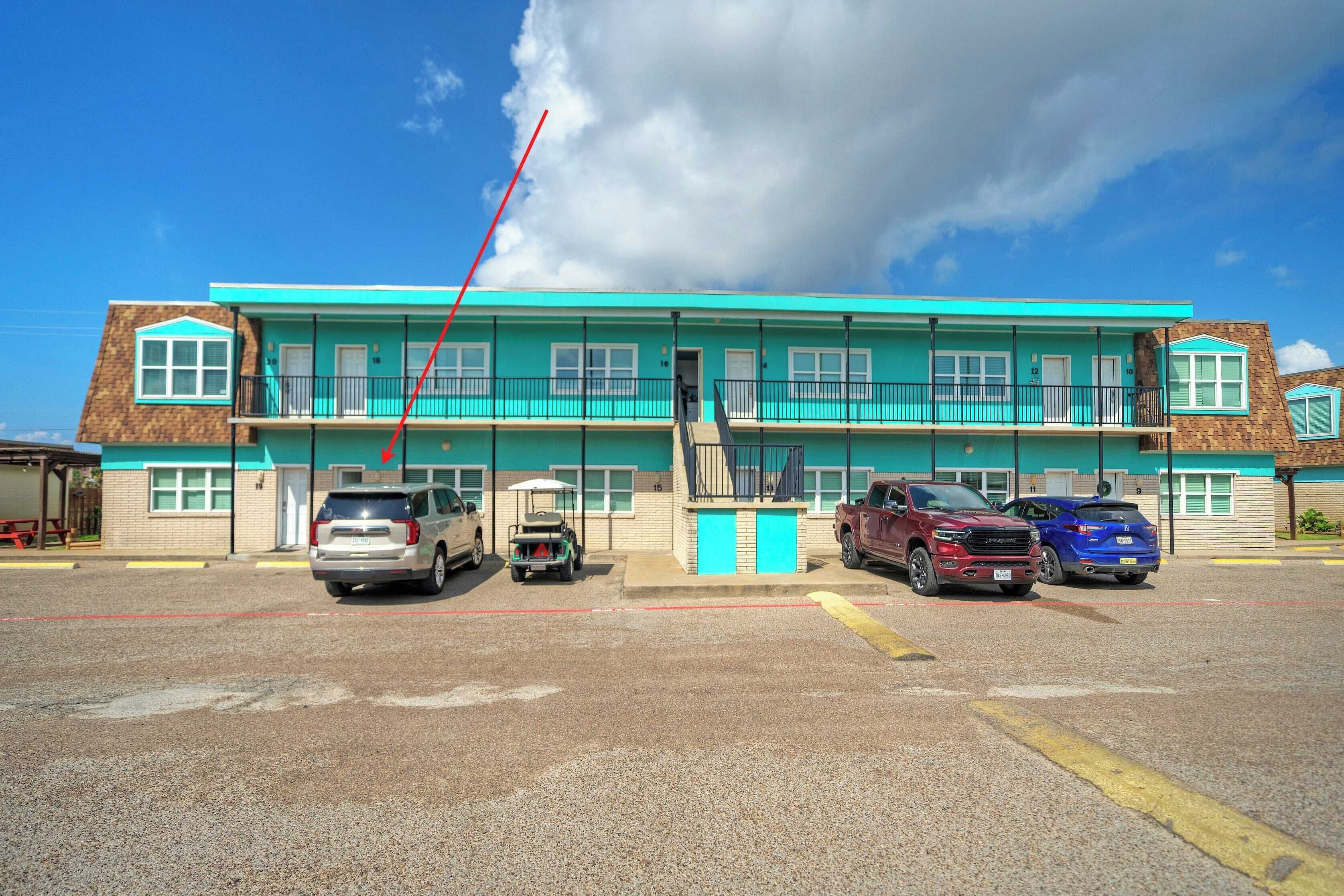 Un bâtiment à deux étages doté d’une façade turquoise, d’un escalier central et d’un parking où sont stationnés plusieurs véhicules.