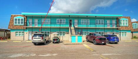 Un bâtiment à deux étages doté d’une façade turquoise, d’un escalier central et d’un parking où sont stationnés plusieurs véhicules.