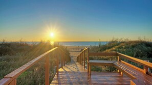 Ein Holzsteg führt zu einem Strand, im Hintergrund versinkt die Sonne.
