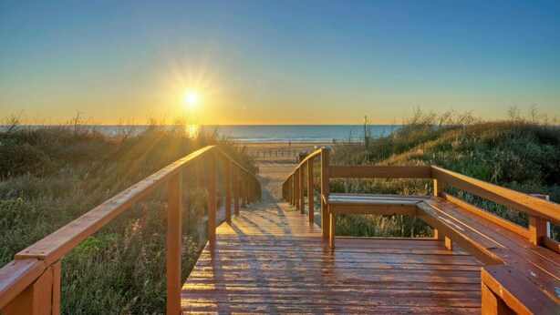 Une promenade en bois qui mène à une plage, avec le soleil couchant en arrière-plan.