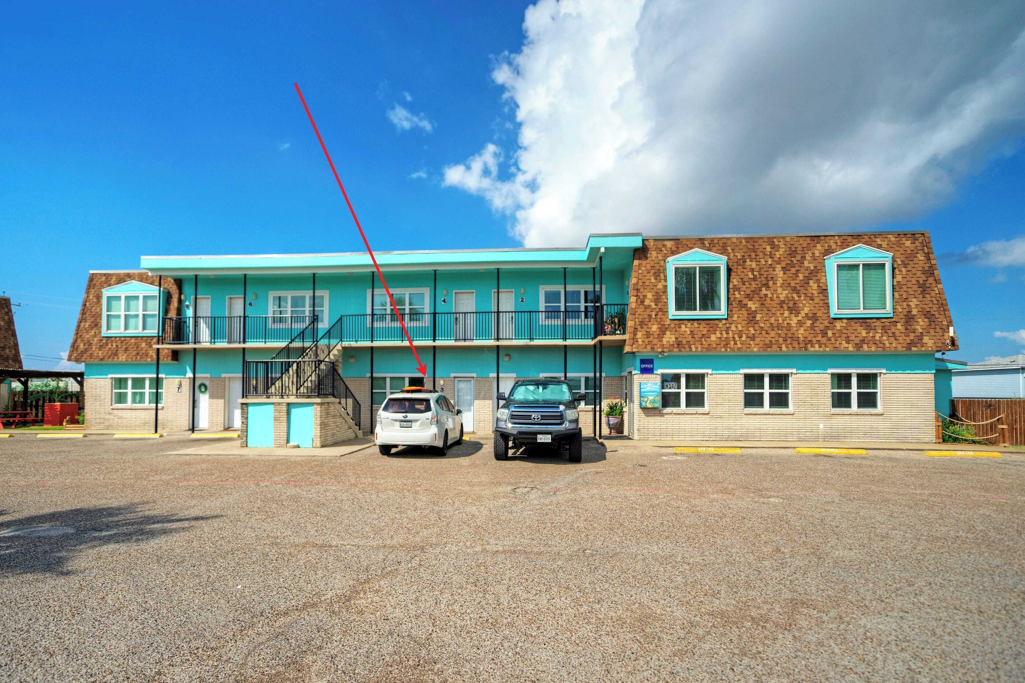 Et motel med en turkis facade, to etager og en parkeringsplads med grus.