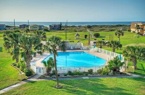 Una piscina rodeada de palmeras y una valla blanca, con vista al océano y casas de playa al fondo.