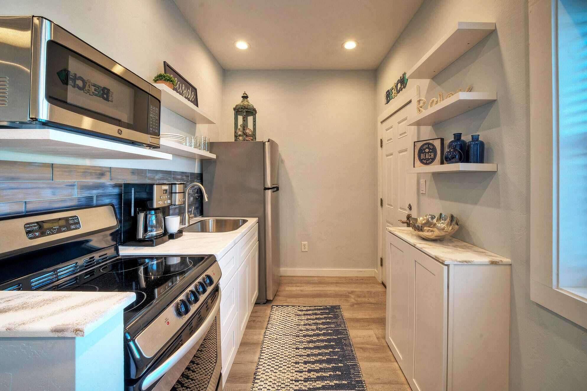 Cocina compacta con estufa, microondas y refrigerador. Hay repisas con objetos decorativos y una alfombra pequeña en el suelo.