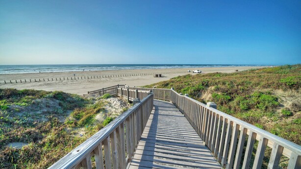Ein hölzerner Steg führt zu einem Strand unter blauem Himmel.