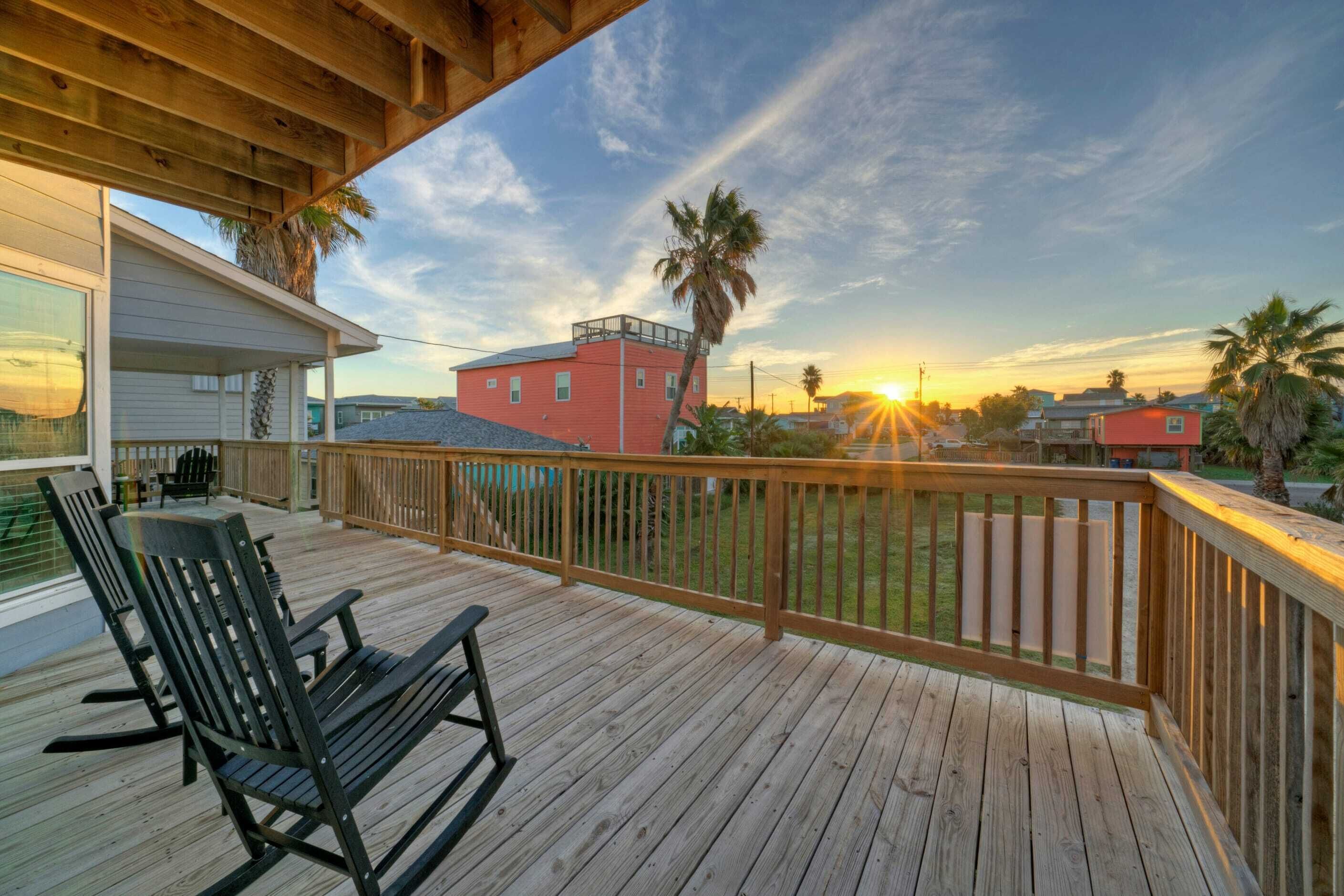 A wooden deck with rocking chairs, a view of a sunset, and a red building in the background.
