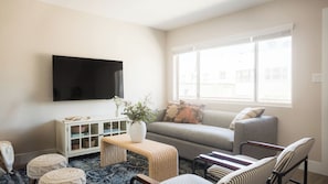 A living room with a flat-screen TV, a sofa, a window with blinds, a wicker bench, and a striped chair.