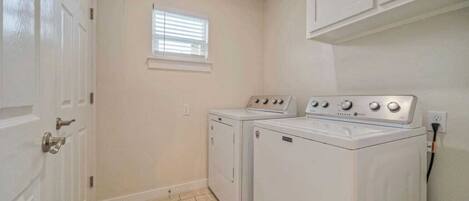 A laundry room with two washing machines and a dryer, a window with blinds, and a door.