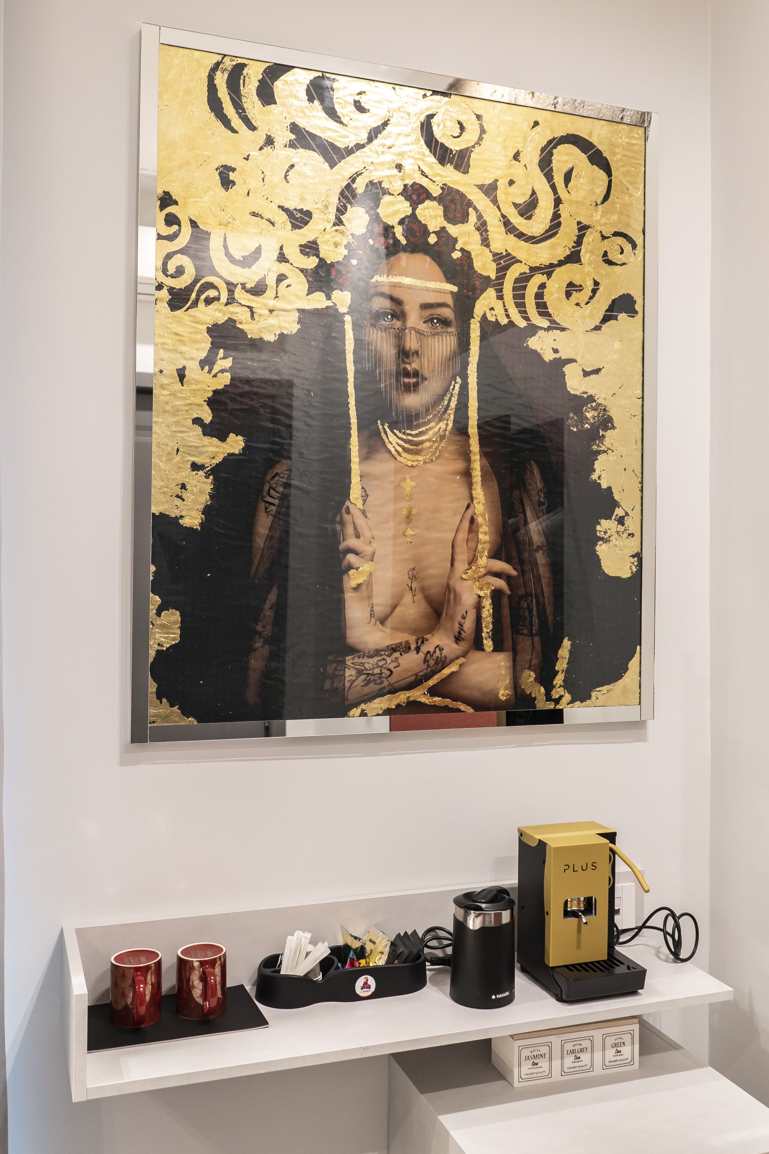 A framed artwork with a gold and black design, a coffee machine, and two red mugs on a white shelf.
