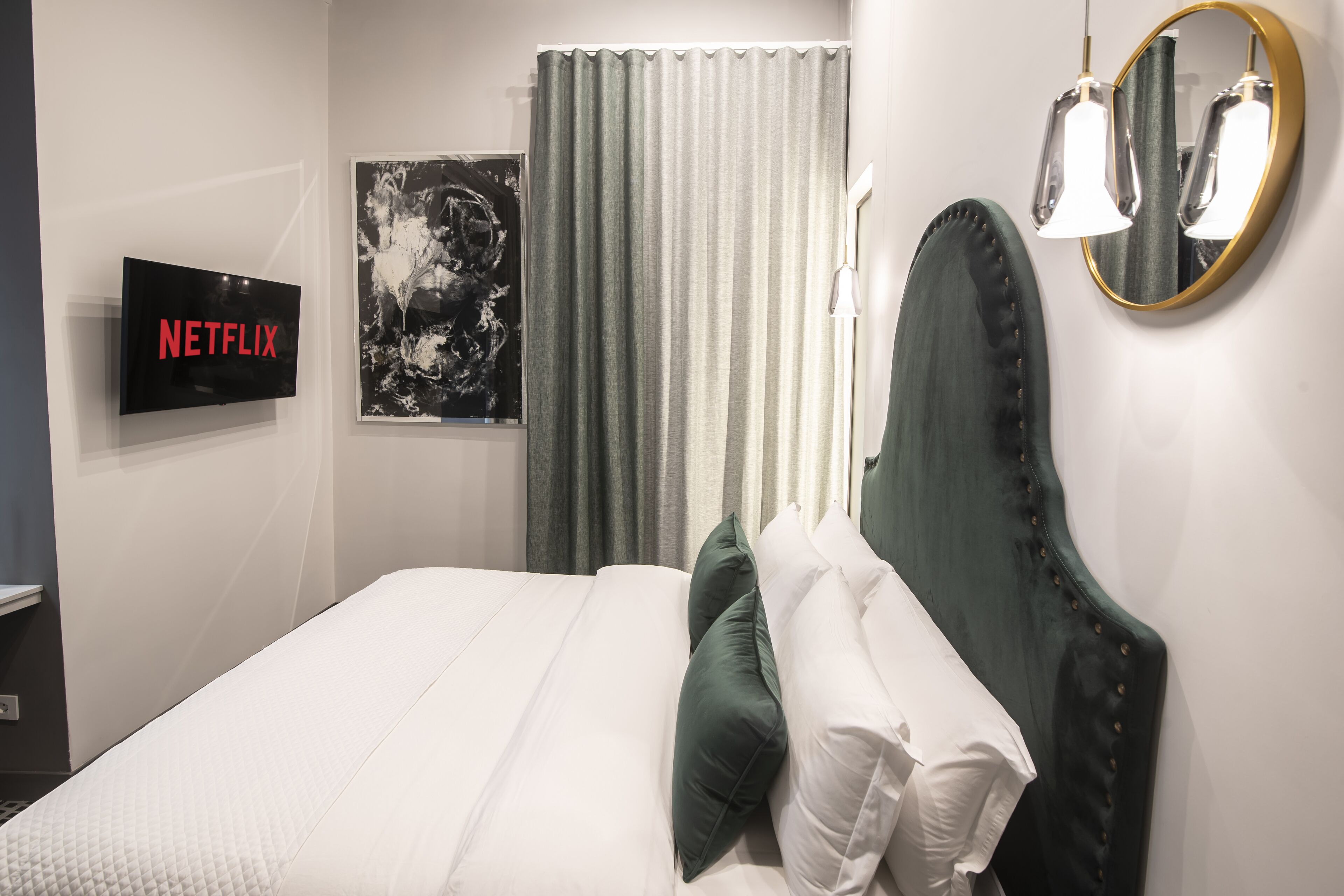 A neatly made bed with white and green pillows, a mounted TV displaying 'NETFLIX', a framed abstract artwork, and a mirror with a gold frame.