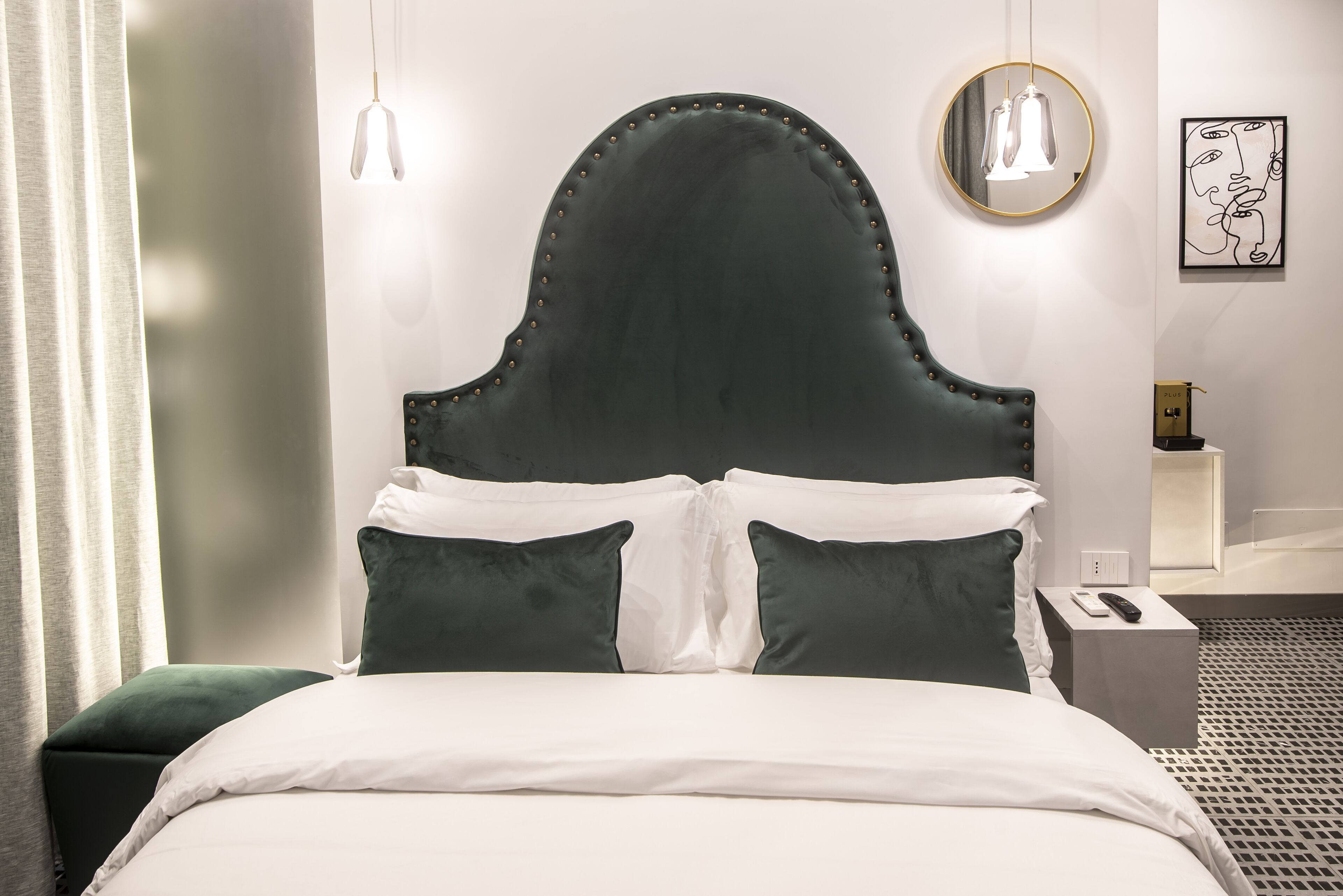 Una cama bien hecha con un cabecero oscuro, ropa de cama blanca y almohadas verdes. Hay una mesita de noche con una lámpara y un control remoto.