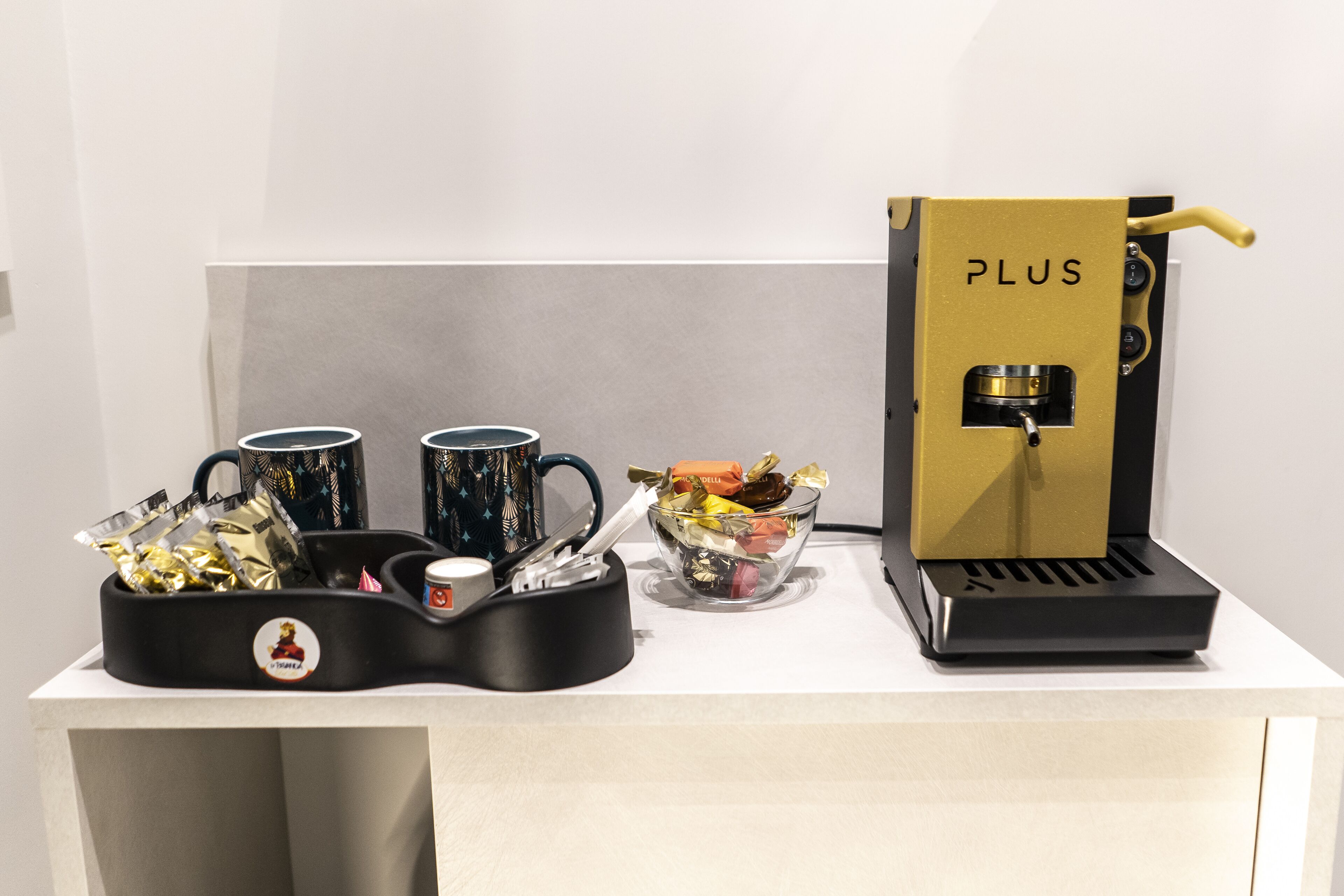 A coffee machine labeled "PLUS" on a white counter with two mugs and a bowl containing various items.