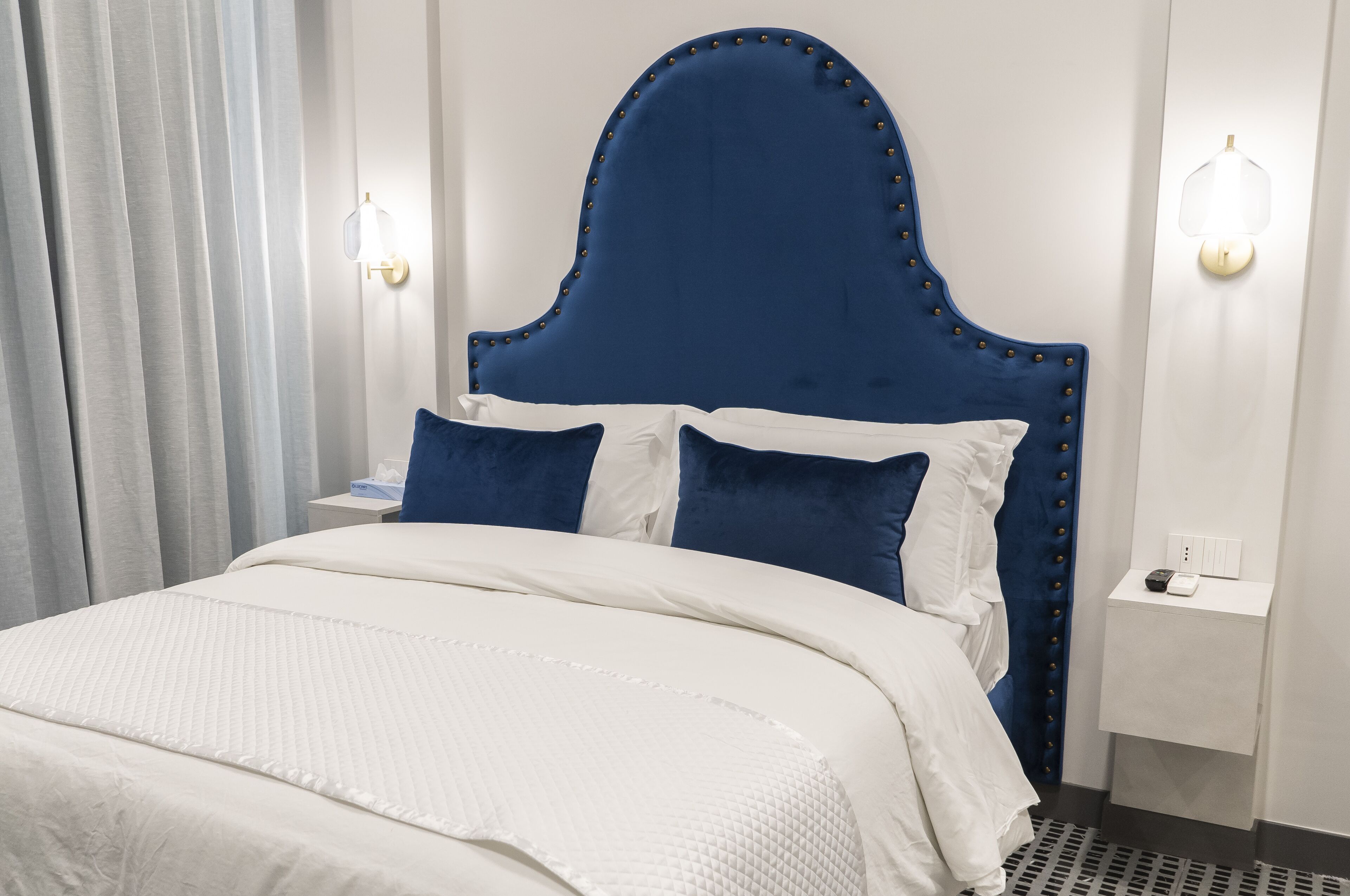 A hotel room with a large bed, white bedding, a blue upholstered headboard, and two wall-mounted light fixtures.
