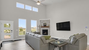 A spacious living room with a grey sectional sofa, a fireplace, a flat-screen TV, and large windows offering a view of the outdoors.