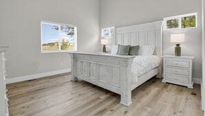 A bedroom with a large bed, two bedside tables, and two windows with views of the outdoors.