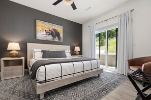 A modern bedroom with a large bed, two bedside tables, a ceiling fan, a large window with curtains, and a chair.