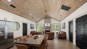 Amplia sala de estar con techo de madera, chimenea, mesa de comedor con sillas y cocina equipada con refrigerador.