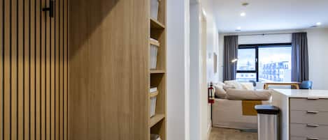 A modern hotel room with a wooden wardrobe, a sofa, a desk, and a view of the city through large windows.