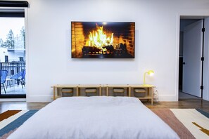 A bedroom with a large bed, a wall-mounted TV displaying a fireplace, a bedside table with a lamp, and a view of the outdoors through a sliding door. - Tumbledown Condo By Revelstoke Vacations (Revelstoke)