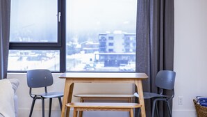 Une pièce comprenant un lit, deux chaises, une petite table en bois et une vue sur des immeubles par la fenêtre.